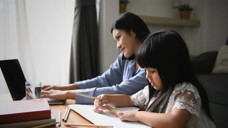 A young Asian mother works from home on a laptop while her child plays next to her.