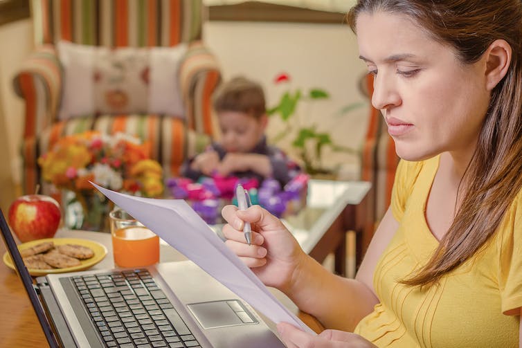 A woman exams documents and works from home while her pre-schooler plays in the background. Half-eaten food and mess sits near her computer.